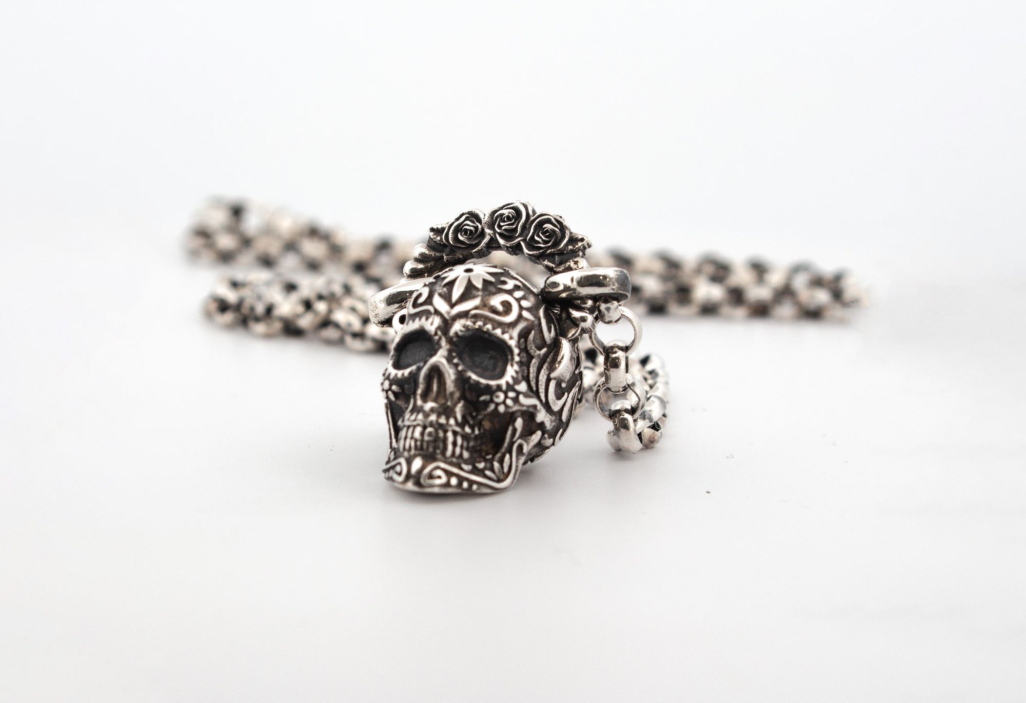 Silver skull pendant with intricate designs and rose details, attached to a beaded chain, against a plain white background.
