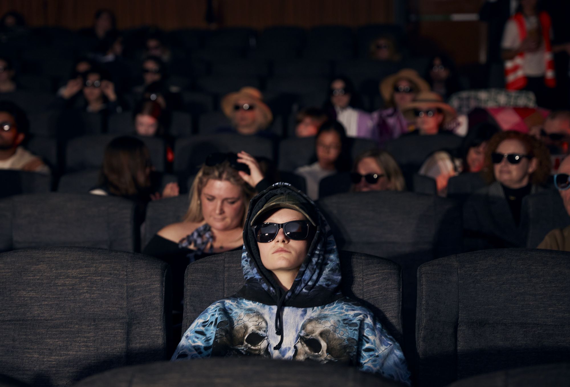 Audience members in a dark cinema, many wearing sunglasses; one person in the foreground wears a hoodie with a skull design and sits with eyes closed.