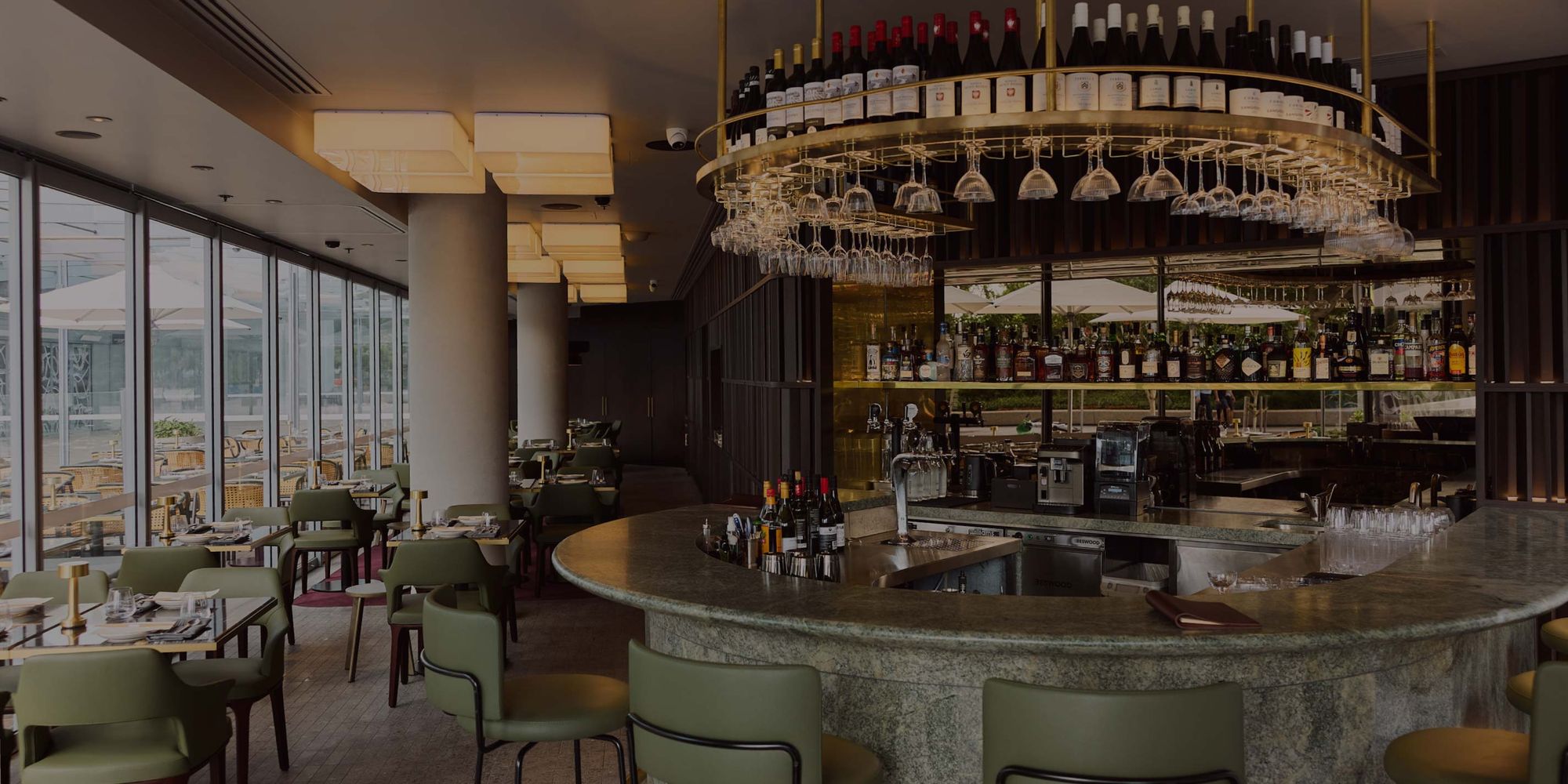 Modern restaurant interior with a central, circular bar, hanging wine glasses, bottles above, and floor-to-ceiling windows offering natural light.