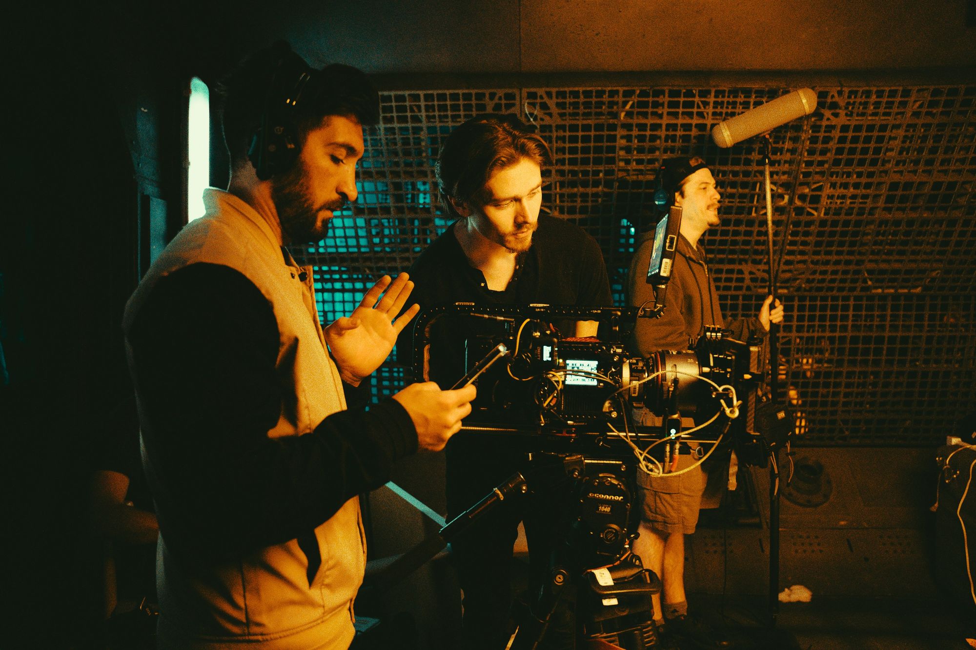 Film crew on set: Two men adjust a camera while another operates a boom mic, in a dimly lit studio with a technical background.