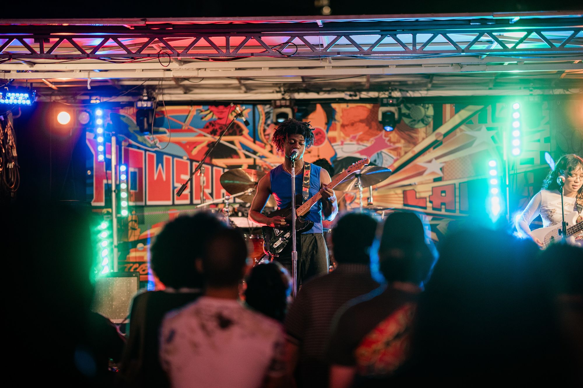 Musician playing guitar and singing on a colorful stage, with an audience watching under vibrant lights.