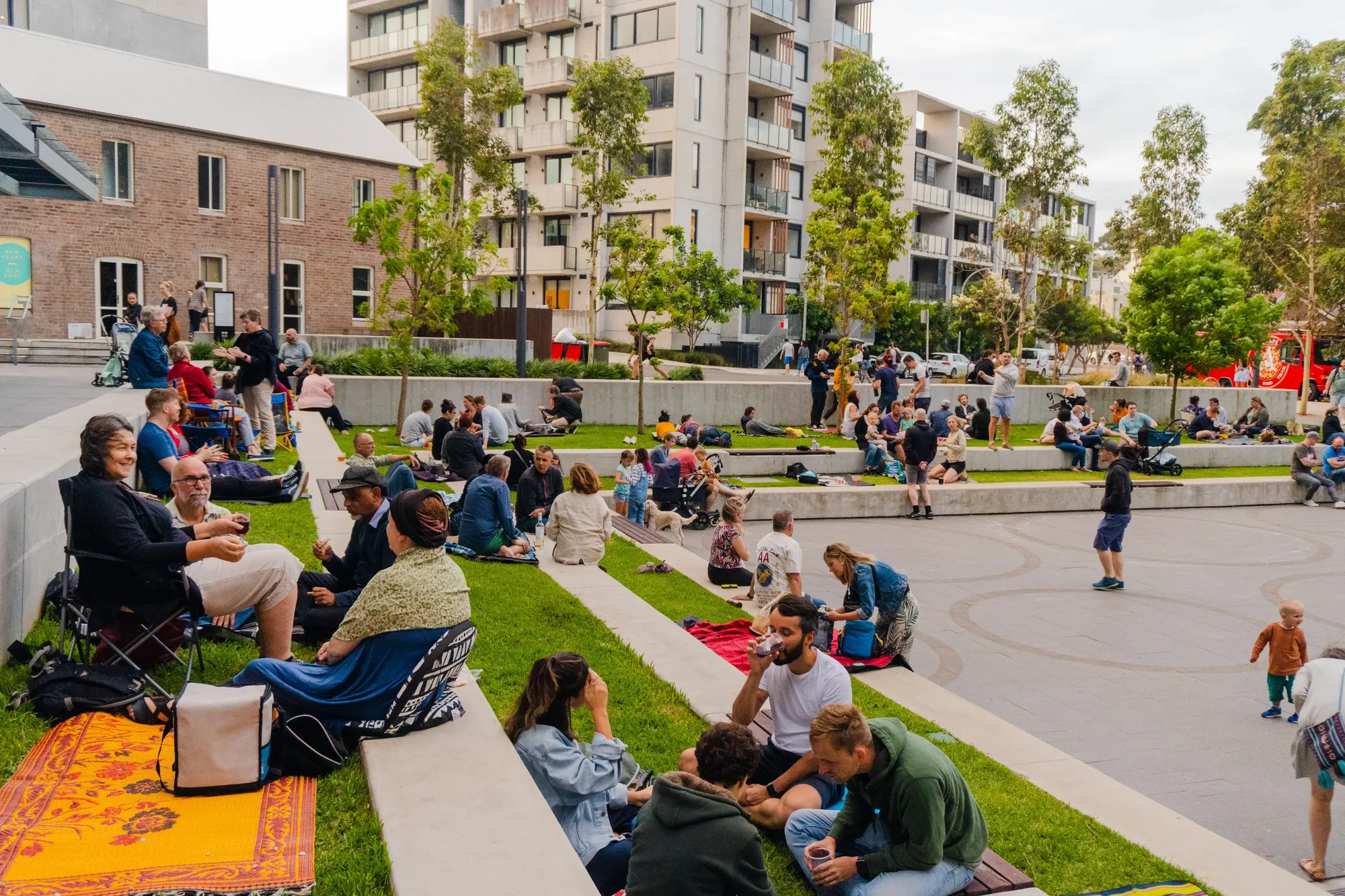 People relaxing on tiered grassy steps in a plaza, surrounded by modern buildings and greenery, with some enjoying picnics and conversations.