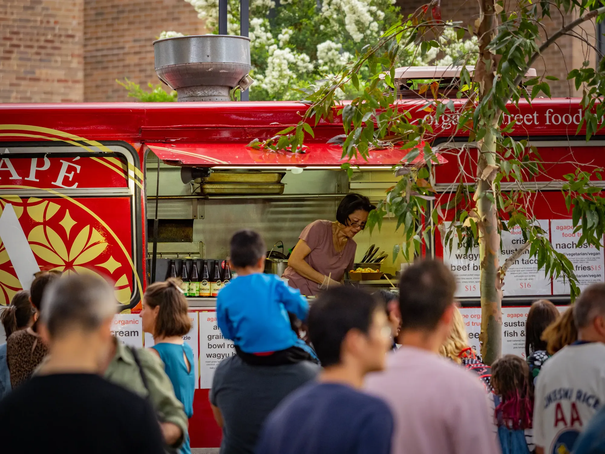 People stand in line at a red food truck with a server inside. A child is on an adult's shoulders, and trees are visible in the background.