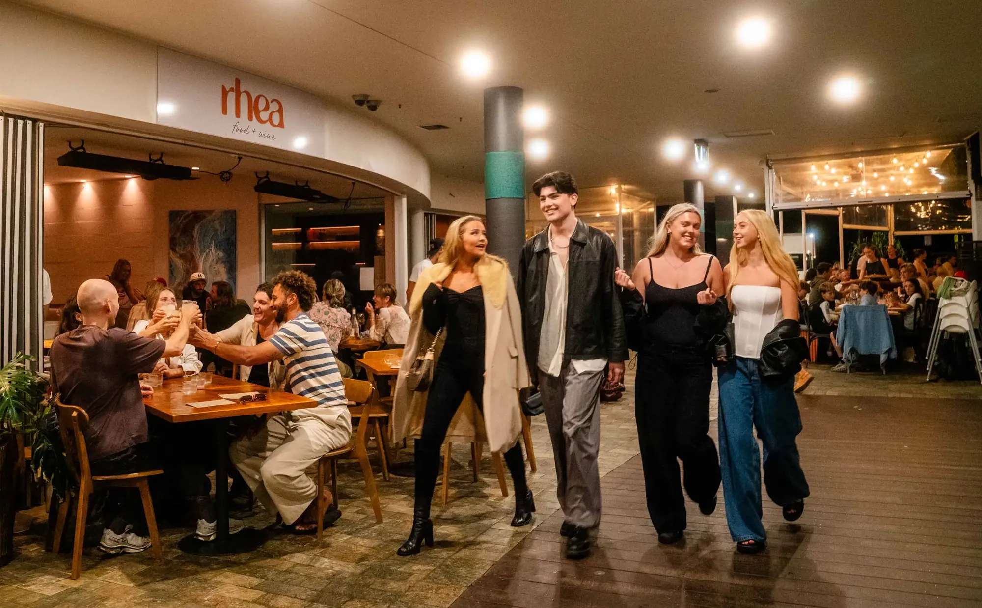A group of people walks past a restaurant, smiling and chatting, while diners inside raise glasses in a toast.