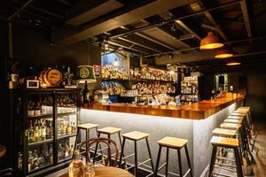 Dimly lit bar with a wooden counter, high stools, a variety of bottles on shelves, and warm ceiling lamps.