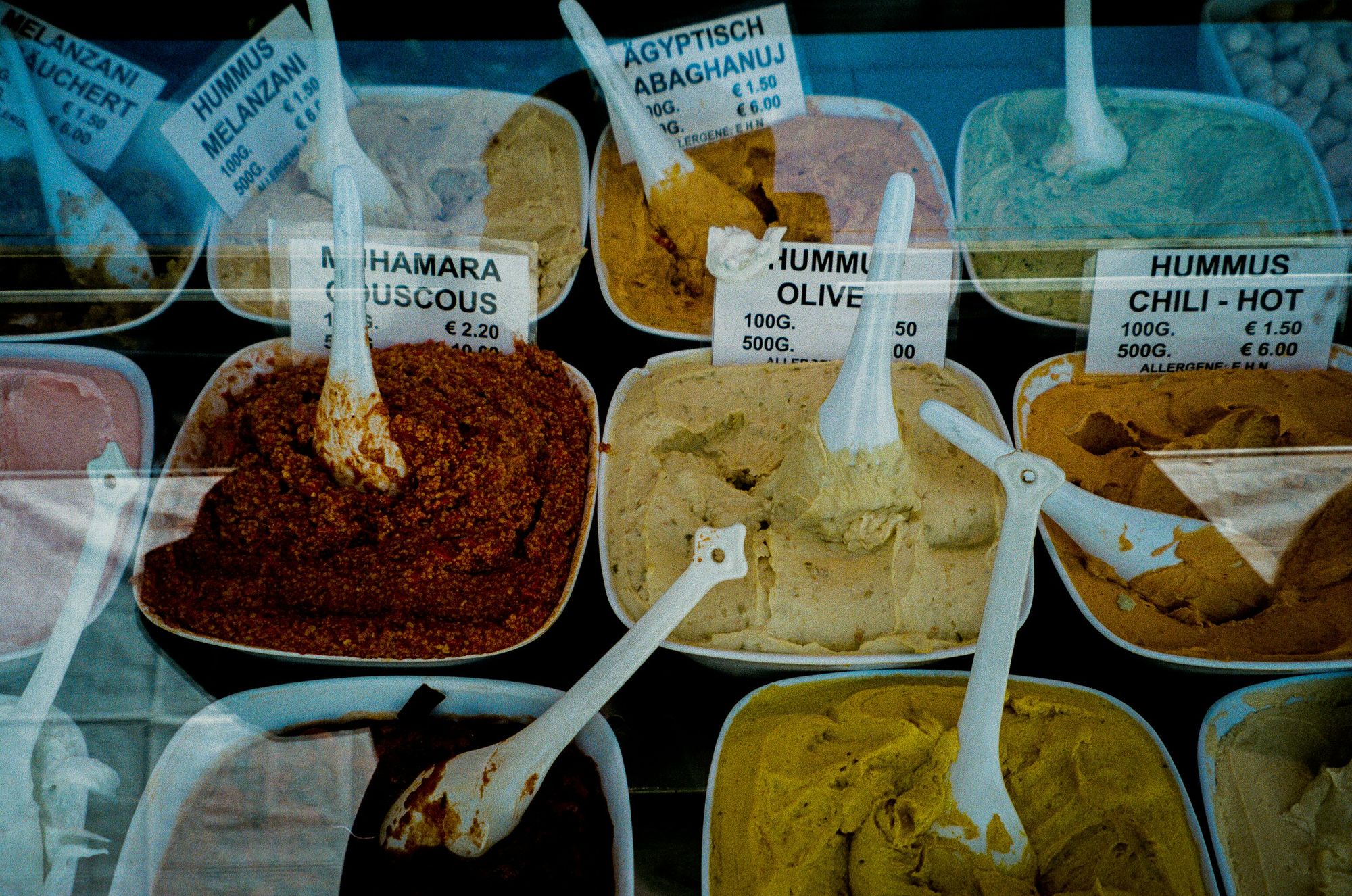 Various hummus and dip varieties with scoops are displayed in containers, each labeled with names and prices, behind a glass counter.