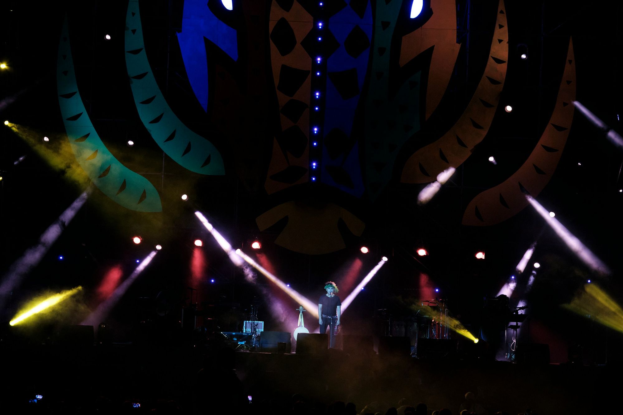 Performer on stage with colorful lights and abstract backdrop, surrounded by beams of light and smoke effects.