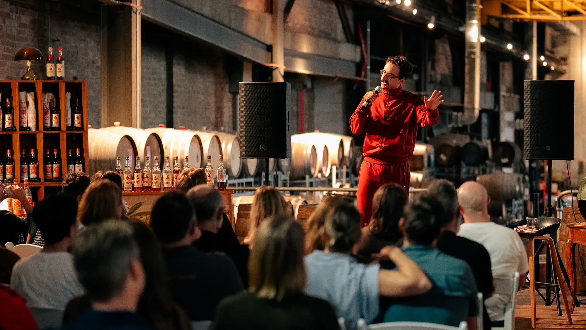 A person in a red outfit holds a microphone, performing to an audience in a room with barrels and bottles on shelves.