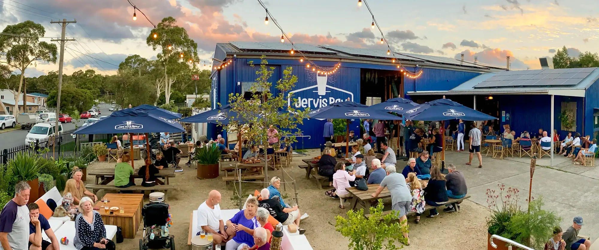 Outdoor dining area with people sitting at picnic tables and under umbrellas at sunset. There are string lights and a building marked "Jervis Bay."
