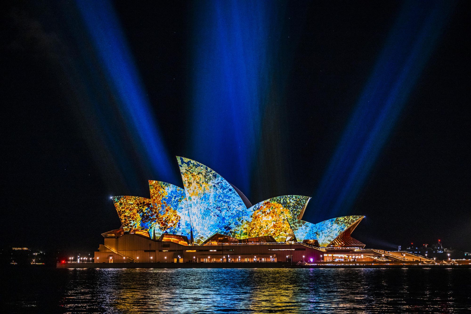 Sydney Opera House illuminated with colourful projections and blue light beams against a night sky, reflecting on the water.
