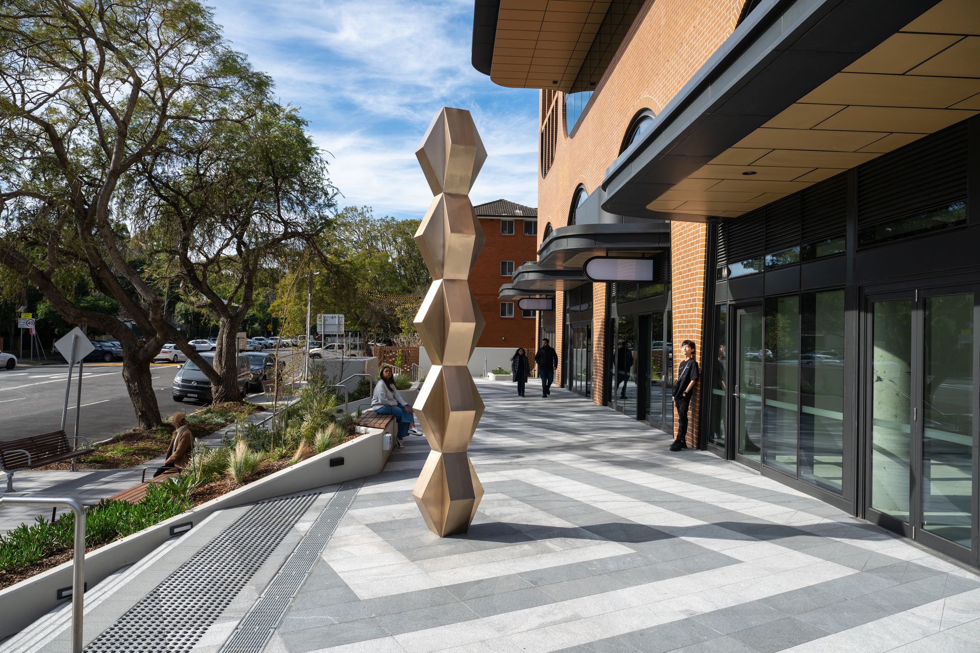 A modern outdoor sculpture of stacked geometric shapes stands on a tiled walkway near a brick building, with people and trees nearby.