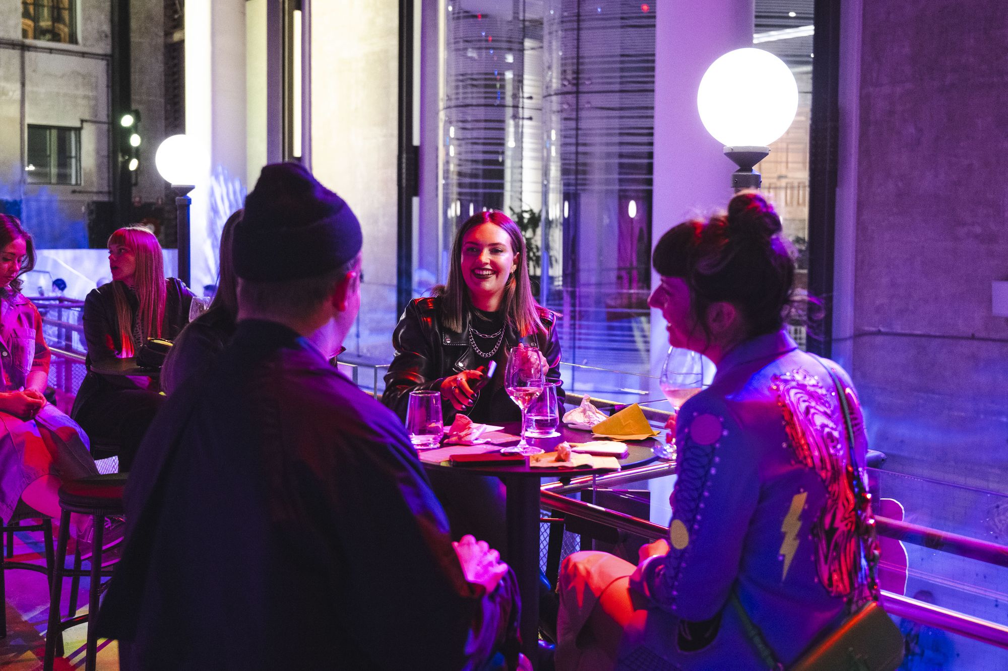 Group of people sitting at a small round table with drinks, chatting and laughing under purple and pink lighting at a Sydney Fringe Festival venue