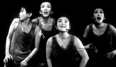 Four performers in expressive poses on stage, wearing dark costumes, with dramatic lighting highlighting their faces against a black background.