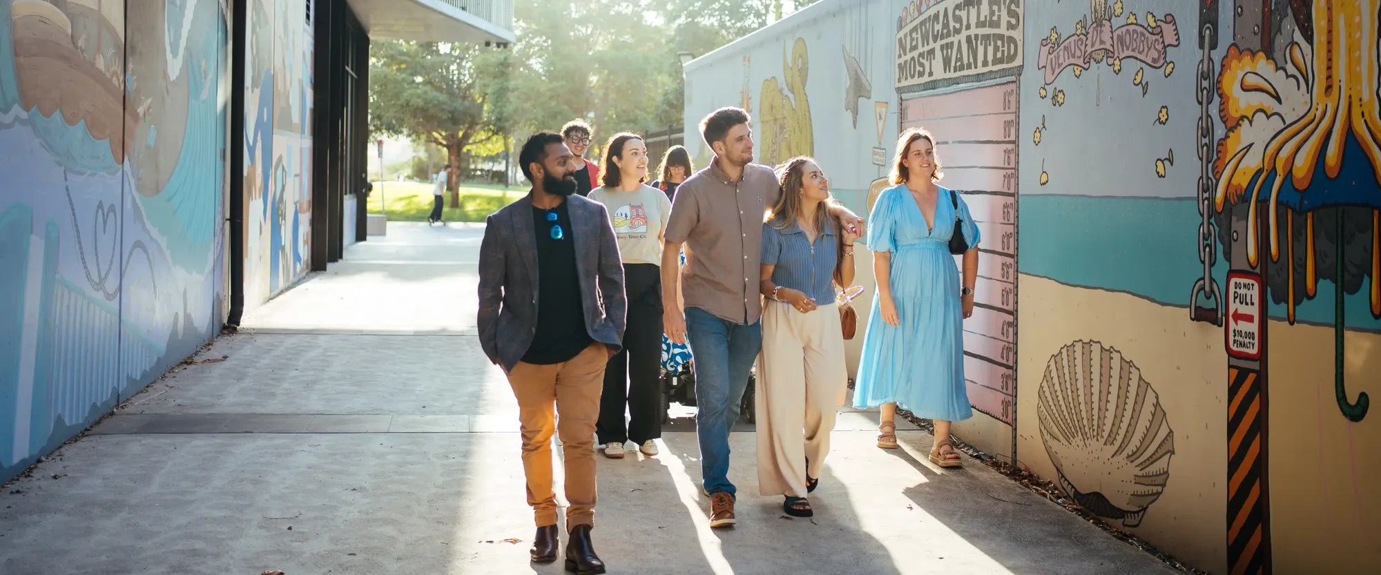 A group of people walking beside a colorful mural on a sunny day, with trees in the background.