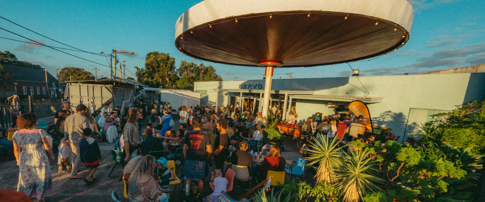 A lively outdoor gathering with people mingling under a large canopy; plants and a bright blue sky in the background.