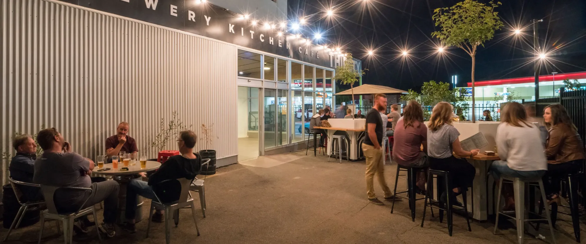 People enjoying an evening at an outdoor seating area of a café, under string lights, with drinks on tables.