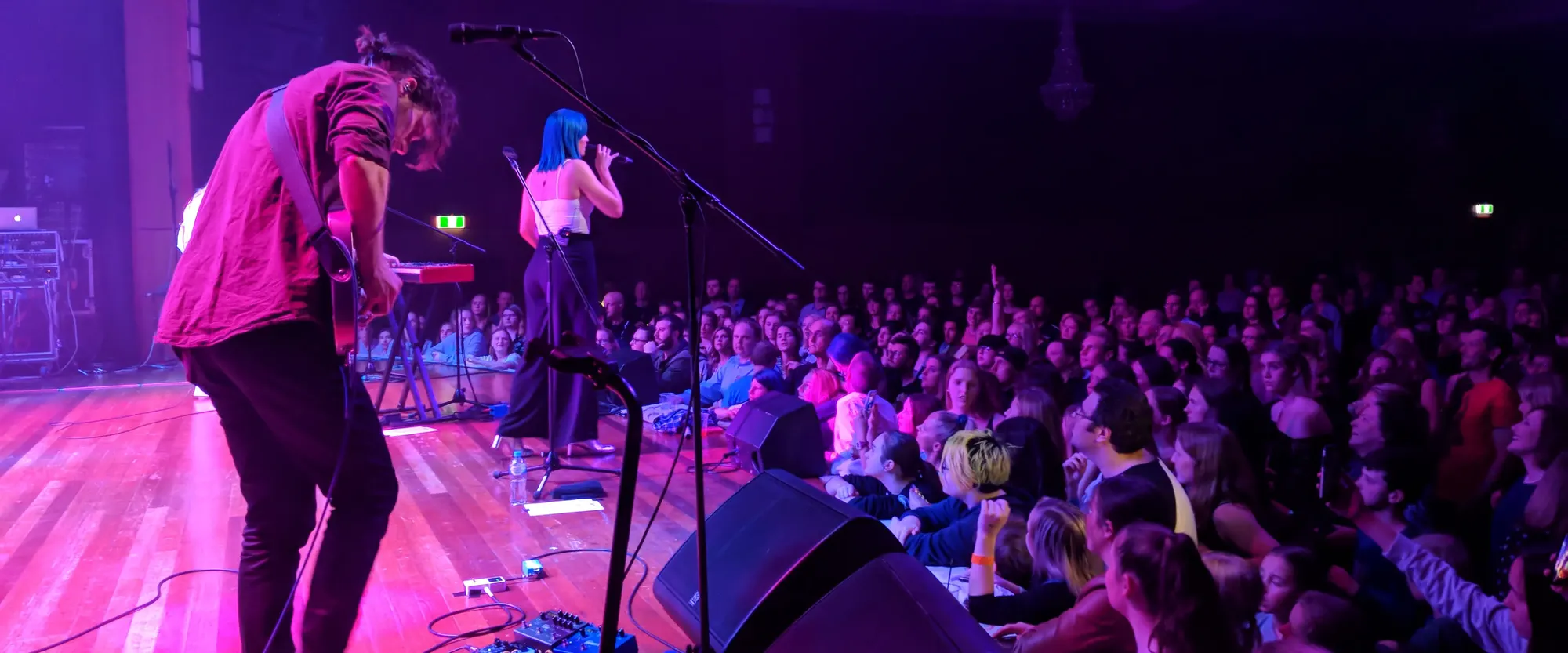 A live concert scene from the stage, showing a guitarist in the foreground, a singer with blue hair performing at center stage, and a large, enthusiastic crowd illuminated by colorful stage lighting.