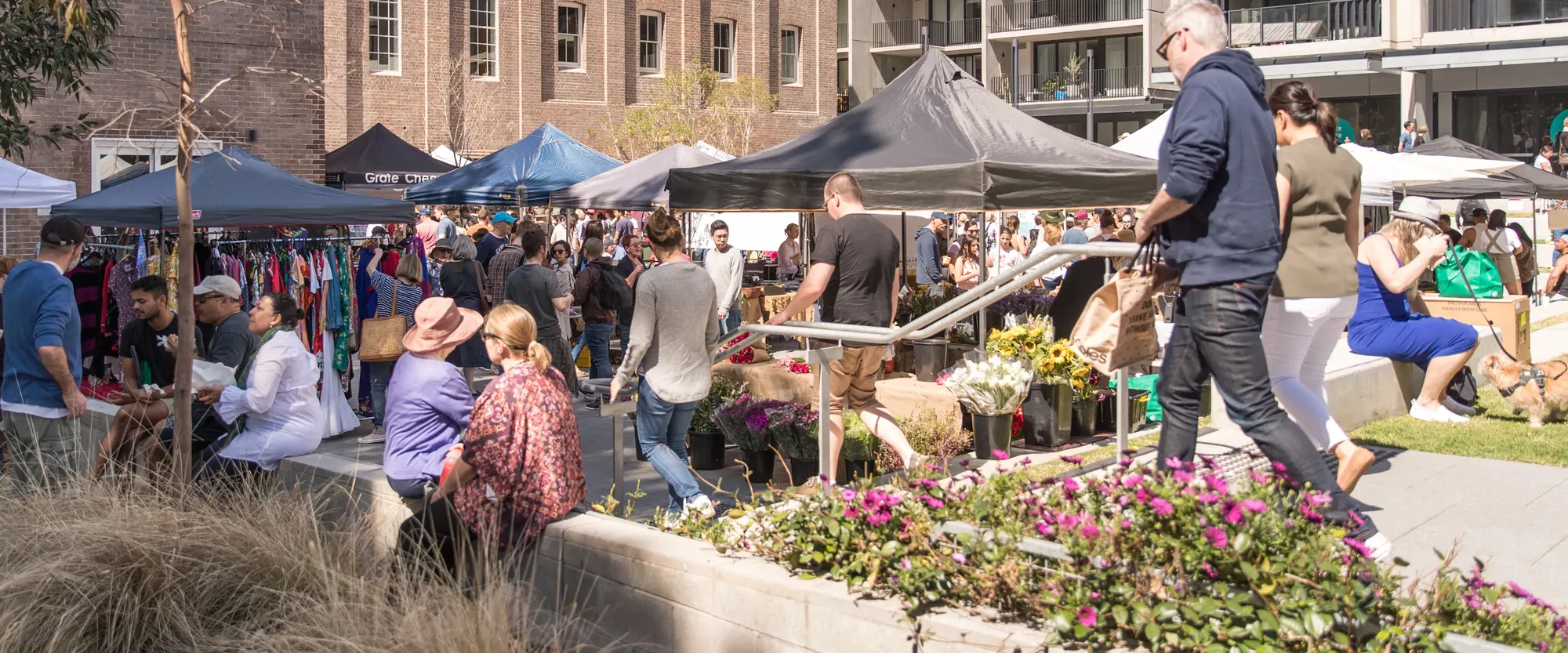 A bustling outdoor market with diverse vendors, flowers, and people mingling under tents in an urban setting.
