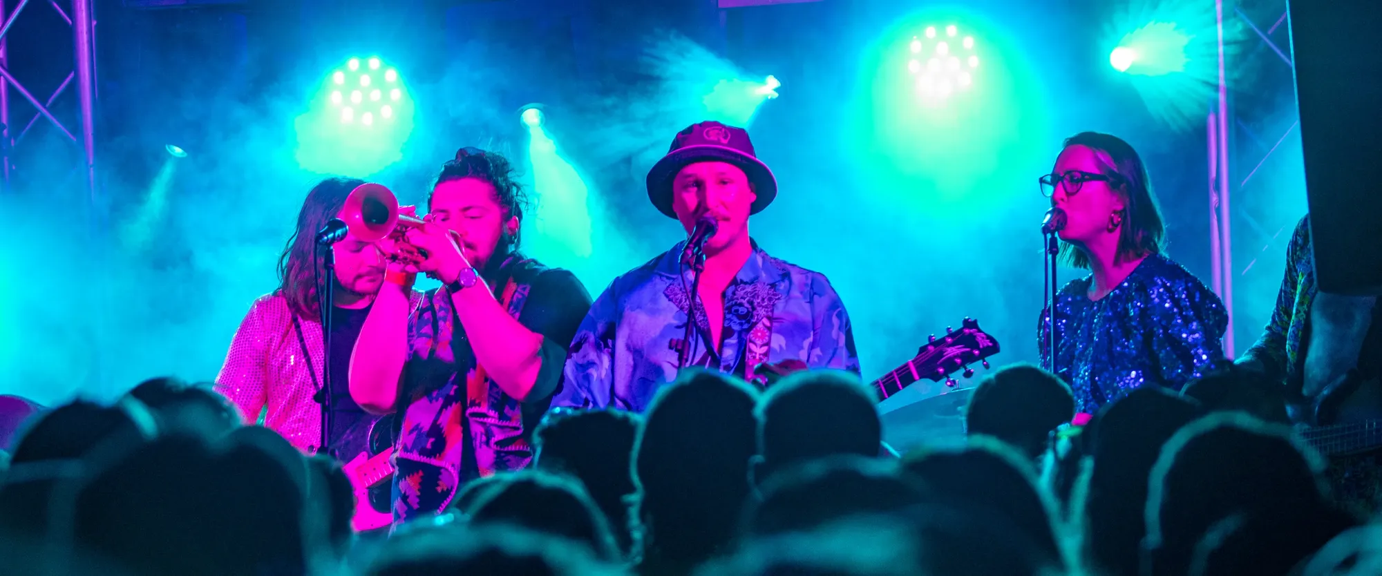 A band performs energetically on stage with vibrant blue and pink lighting, featuring a trumpet player and a guitarist wearing a hat.