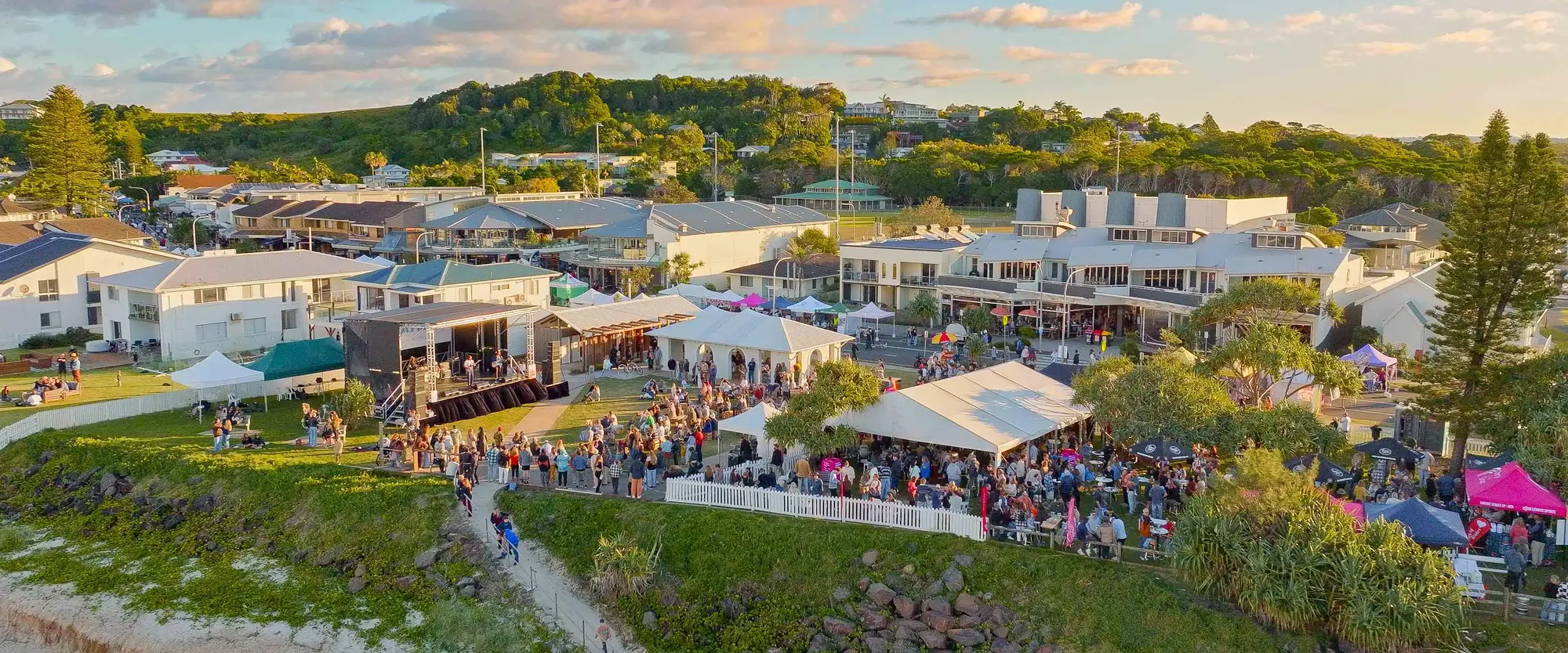 Aerial view of a coastal event with tents, a stage, and a crowd gathered on a grassy area, surrounded by residential buildings and trees.