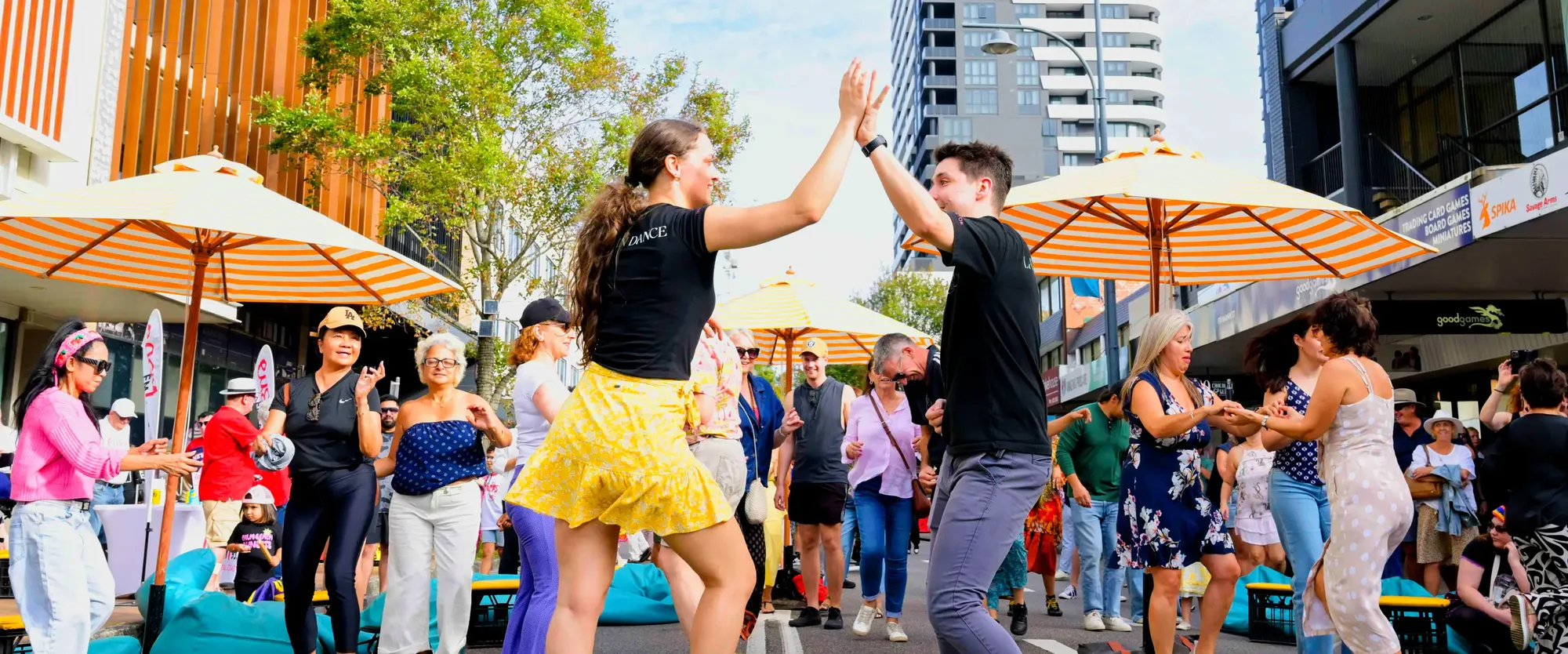 People dancing and socializing outdoors under striped umbrellas, with buildings and trees in the background.
