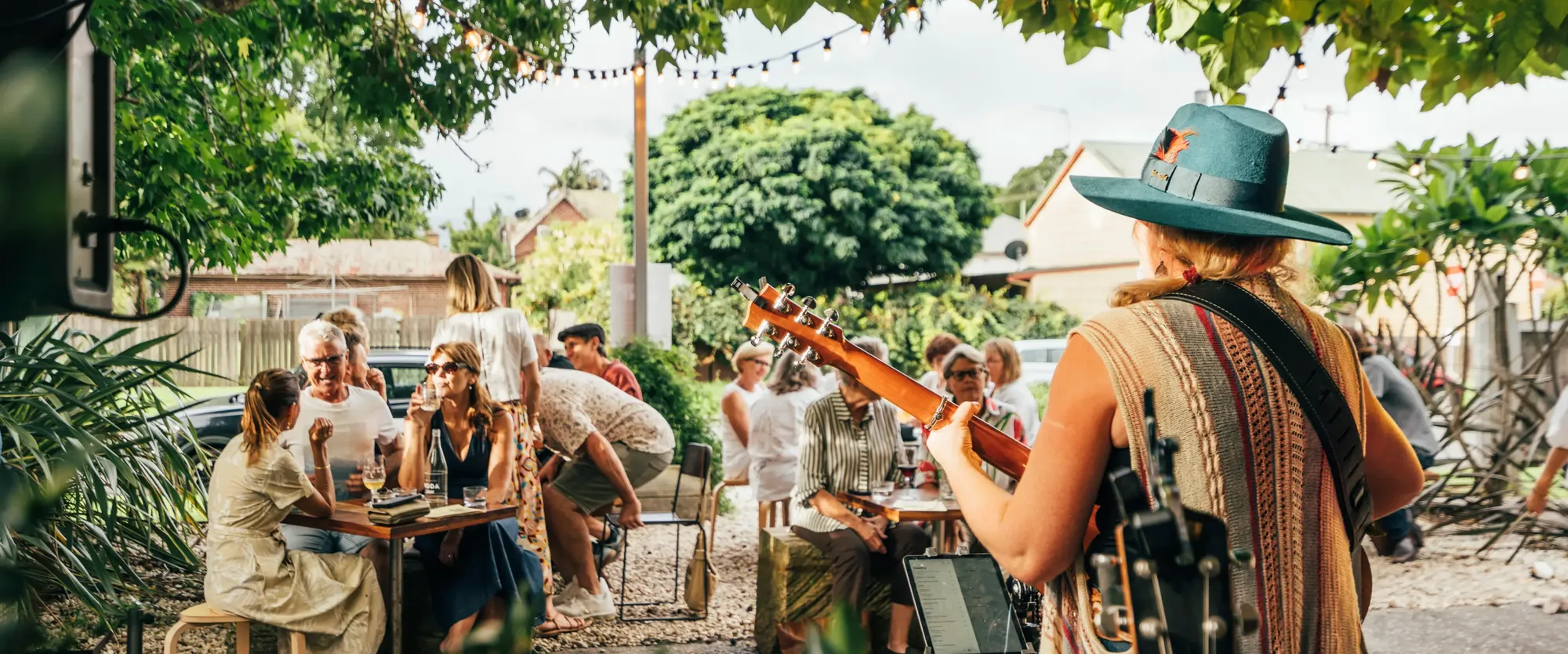 Musician playing guitar at an outdoor gathering, with people sitting at tables under string lights surrounded by greenery.