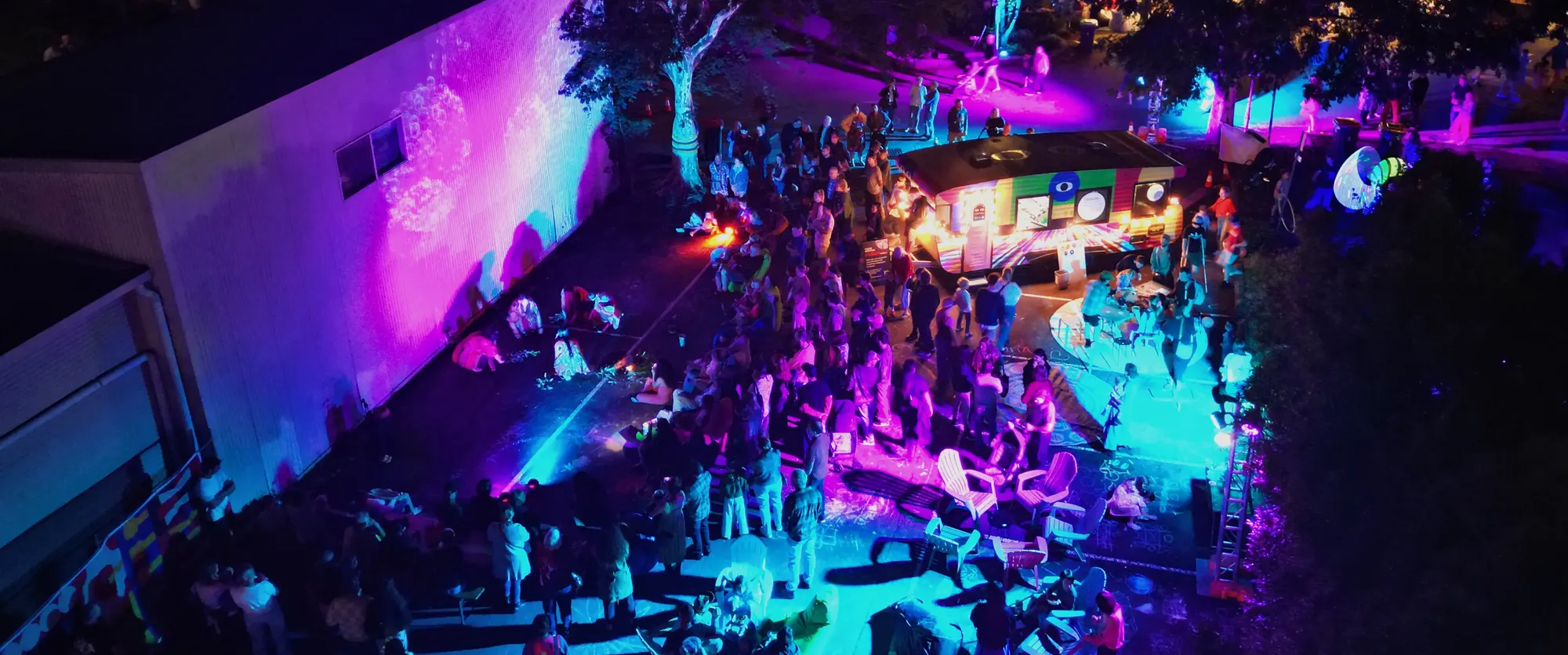 A lively night event with colorful lights, projections, and people gathered around an illuminated food truck and outdoor area.