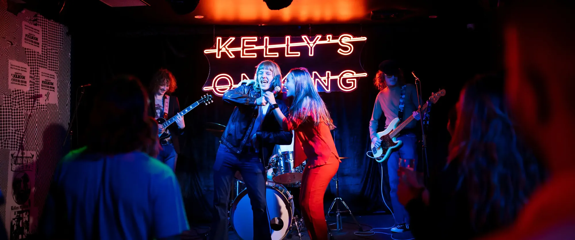 A band performing energetically on stage with neon sign "Kelly's" in the background, as audience members watch and cheer.
