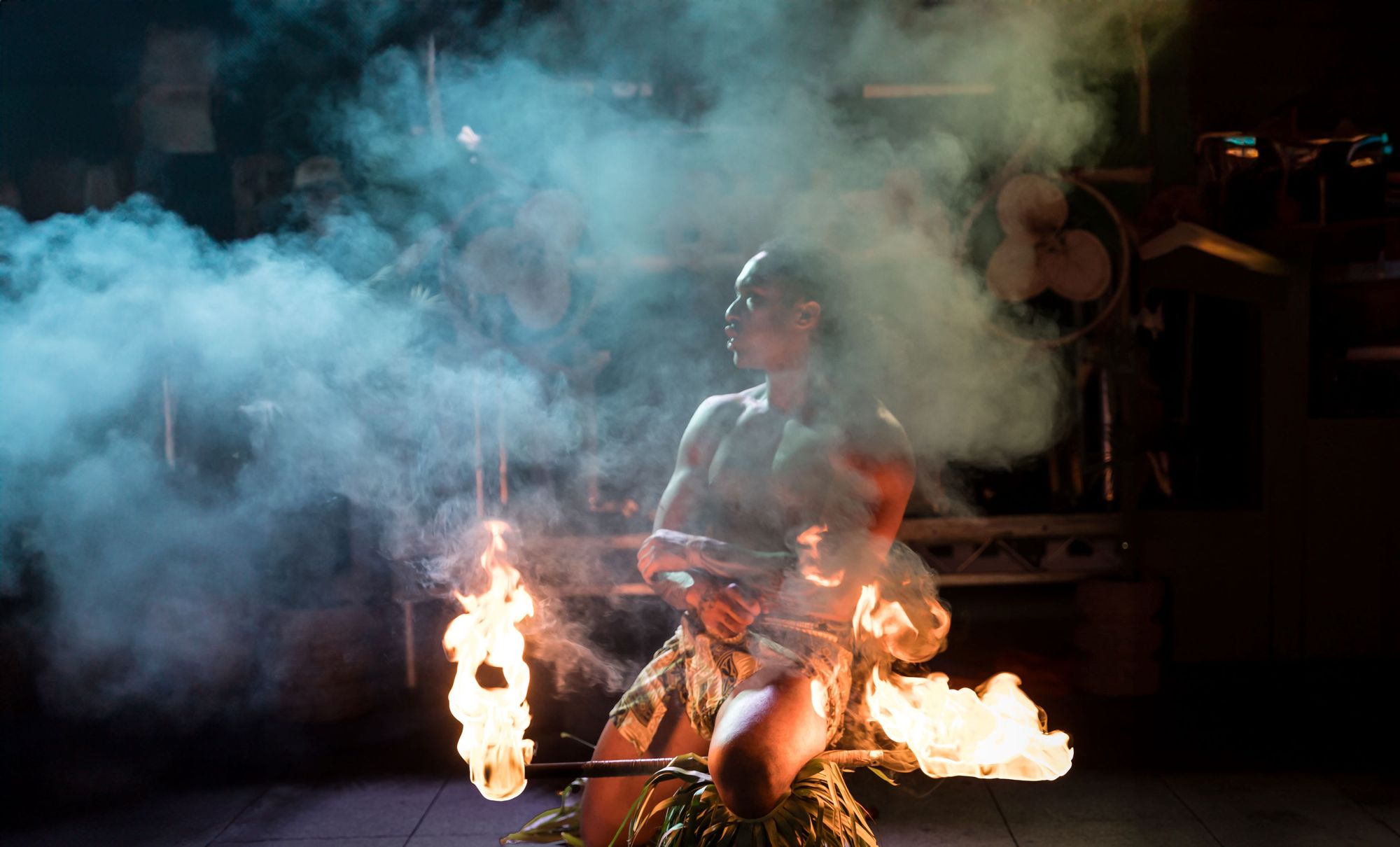 Male fire dancer in traditional dress performing with flaming sticks, surrounded by smoke and dramatic lighting.