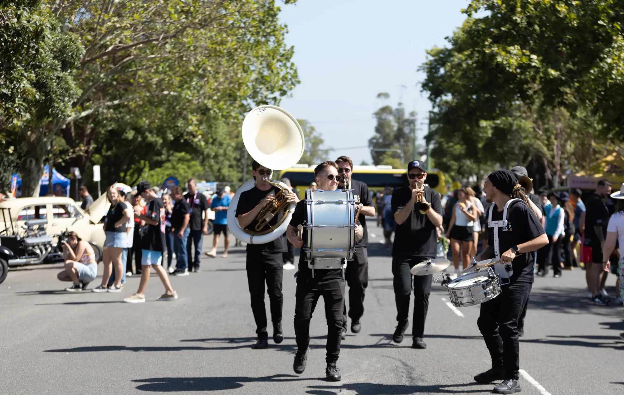 A marching band plays instruments, including a sousaphone and drums, while walking down a street during a sunny outdoor event.