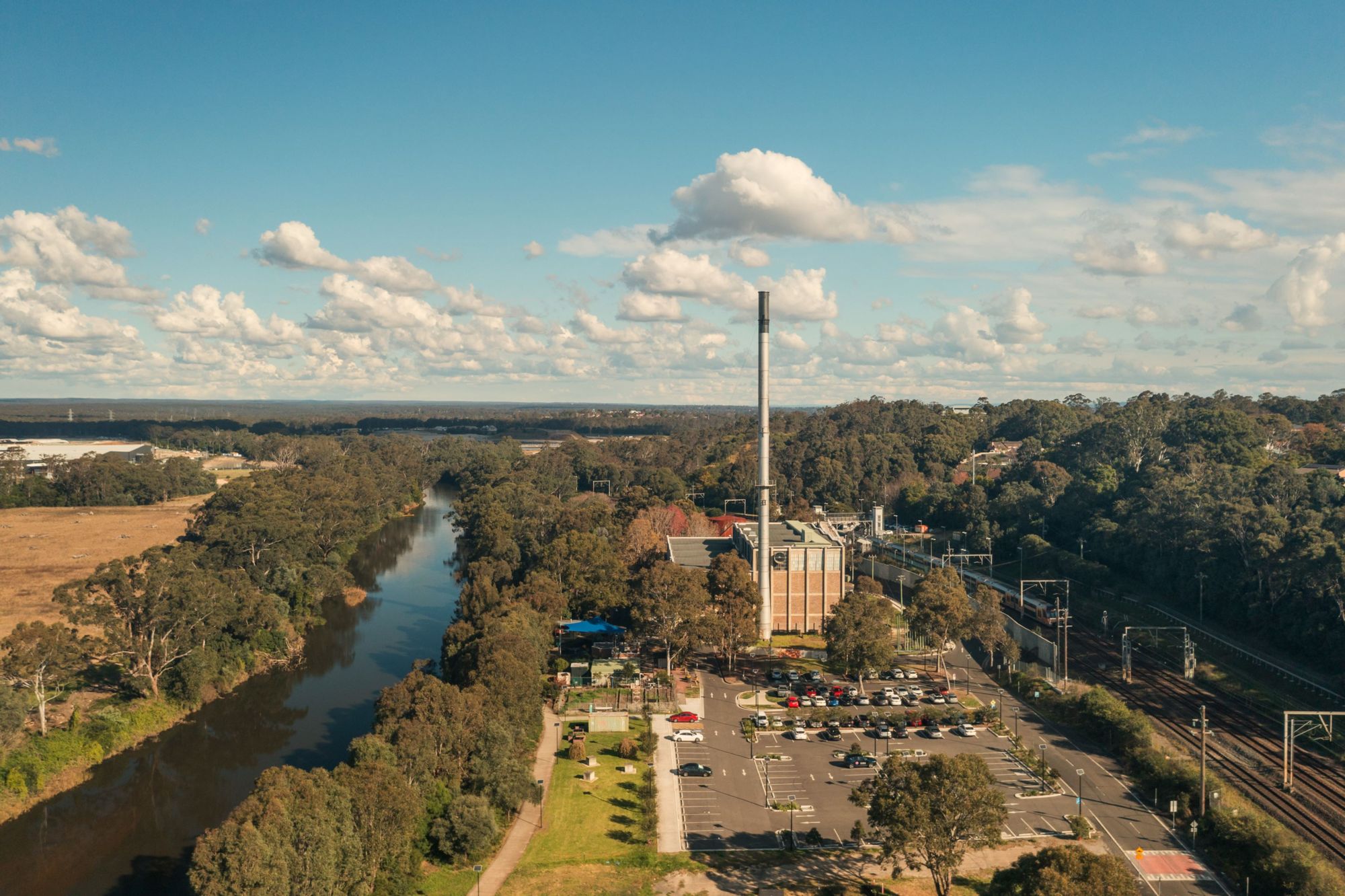 Aerial view of a tall industrial stack next to a river, surrounded by trees and a parking lot.