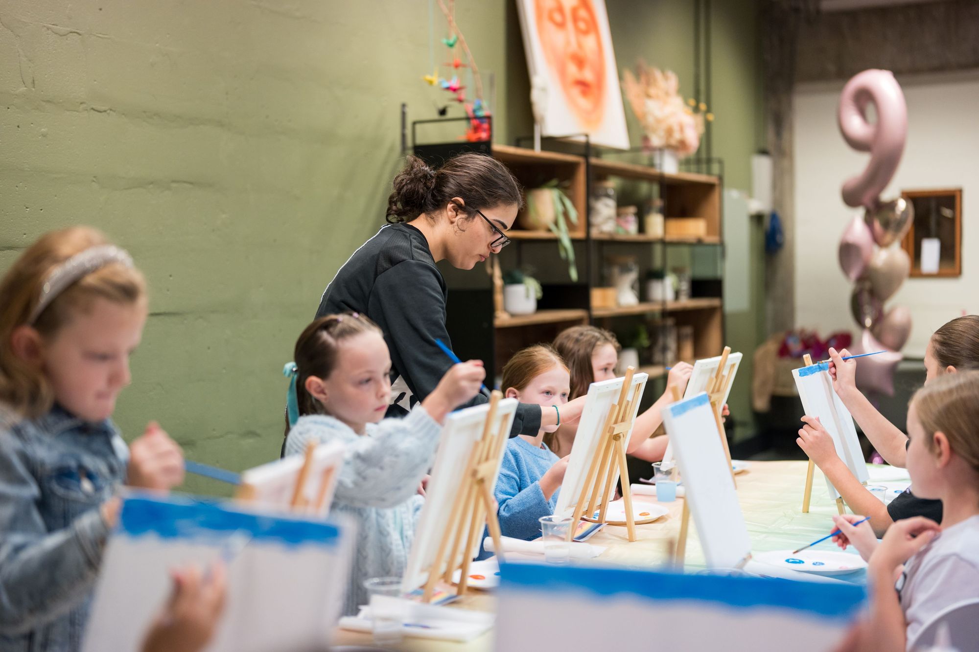 A group of young children are seated at a long table, participating in an art class or paint party. Each child is painting on an easel, and an adult instructor with dark hair is assisting one of the children in the middle of the frame. There are shelves with art supplies and a balloon decoration in the background.