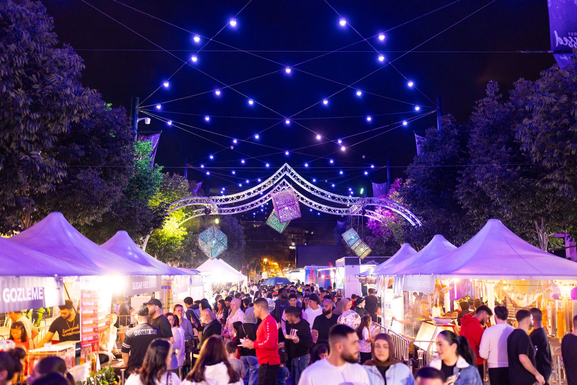 A crowded nighttime food or market festival under blue string lights and illuminated tents.