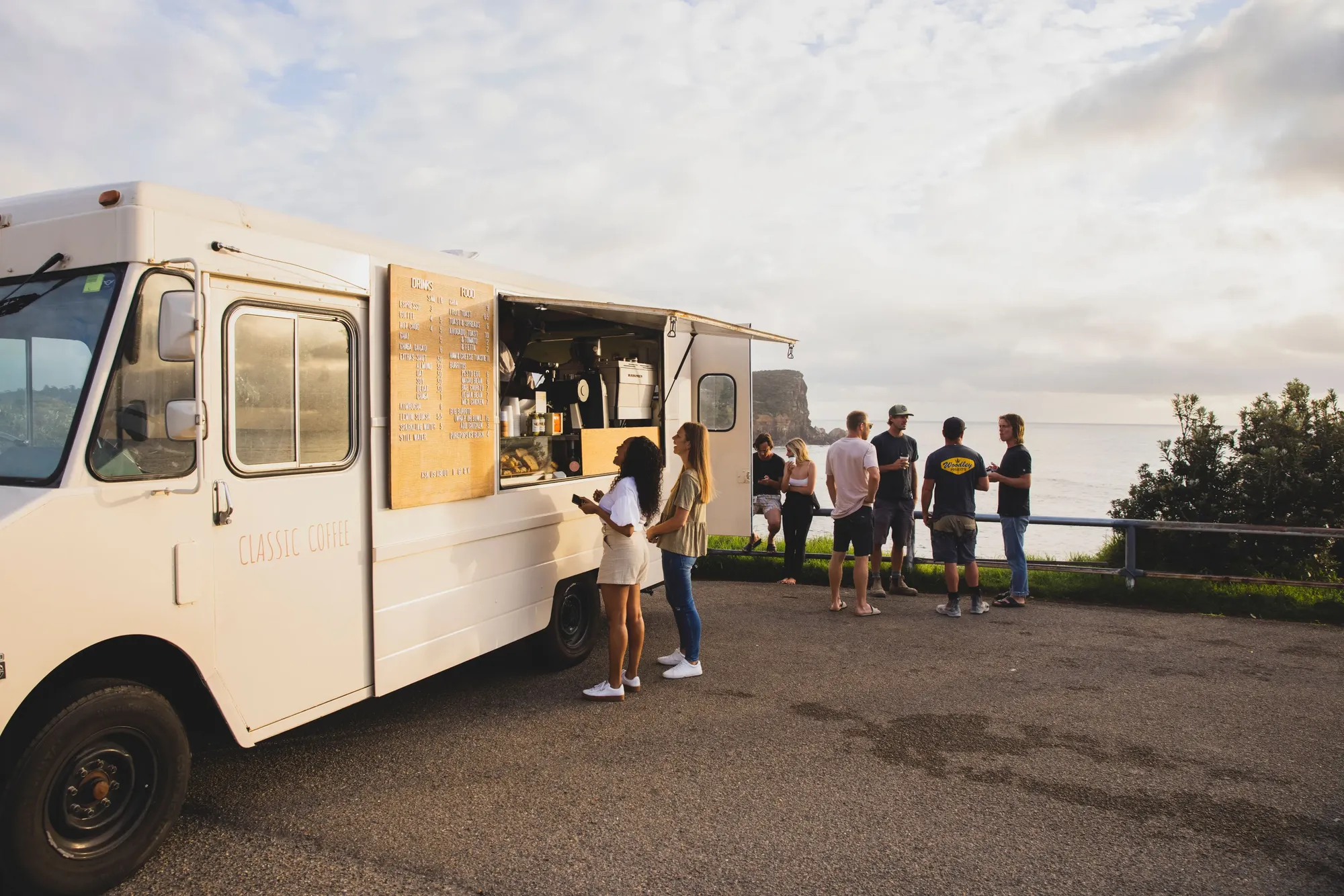 A white food truck serves customers by the seaside at sunset, with several people gathered nearby under a partly cloudy sky.