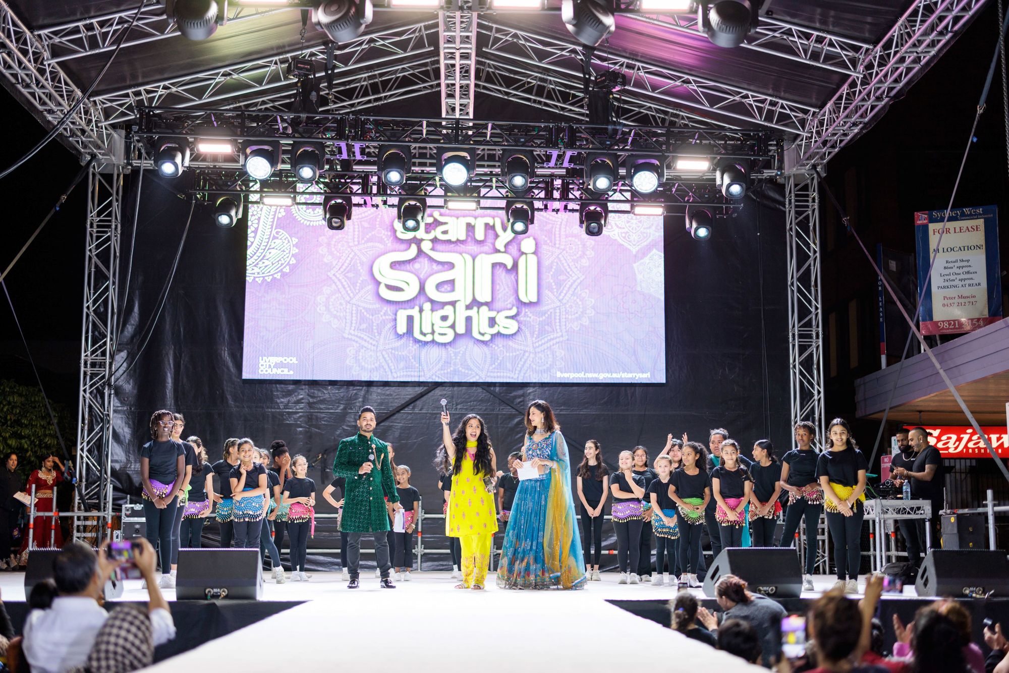Performers in Indian attire singing and dancing on a stage under a screen displaying the Stary Sari Nights logo.