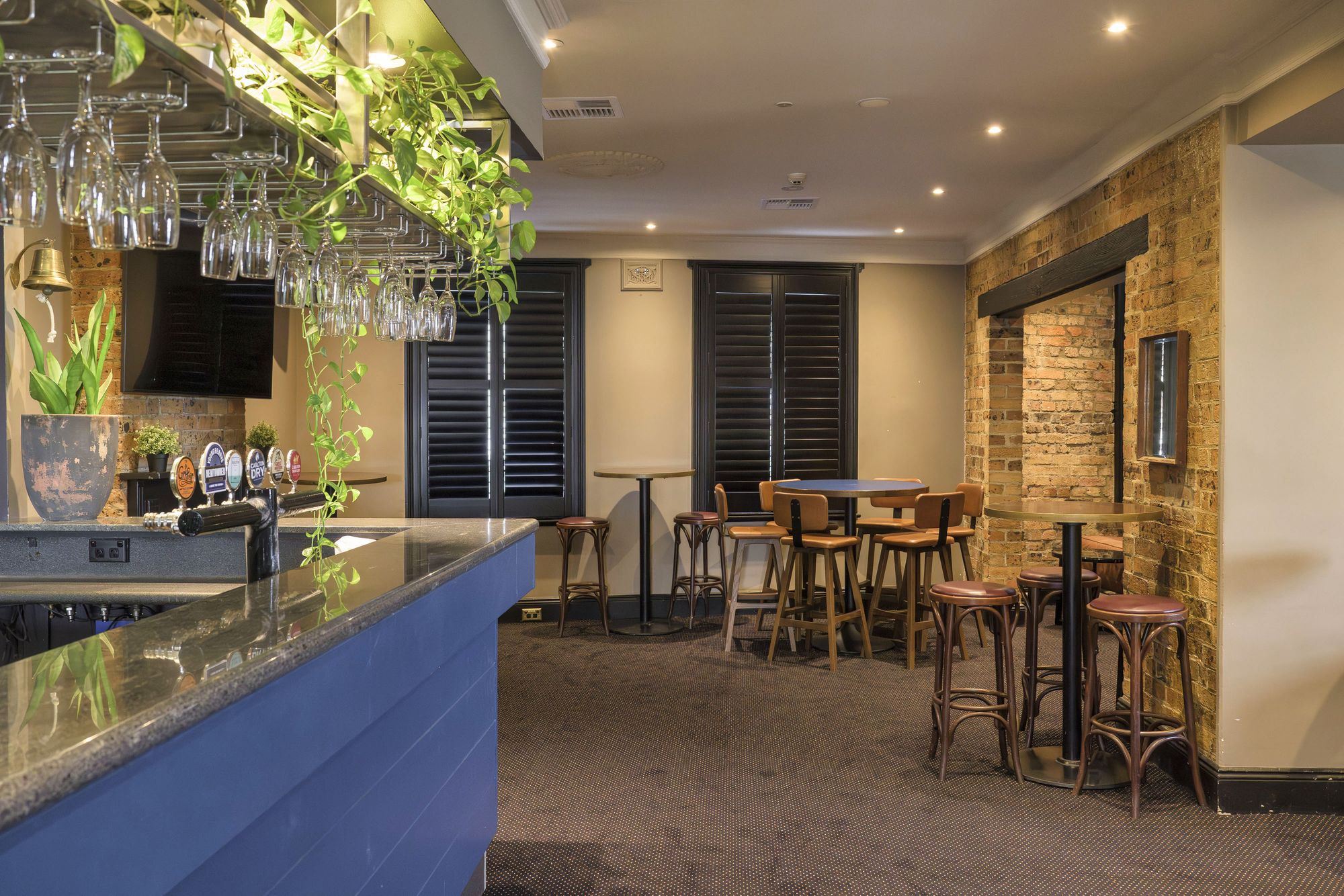 A cozy bar interior with a polished counter, hanging glasses, wooden tables and stools, exposed brick wall, and decorative plants.