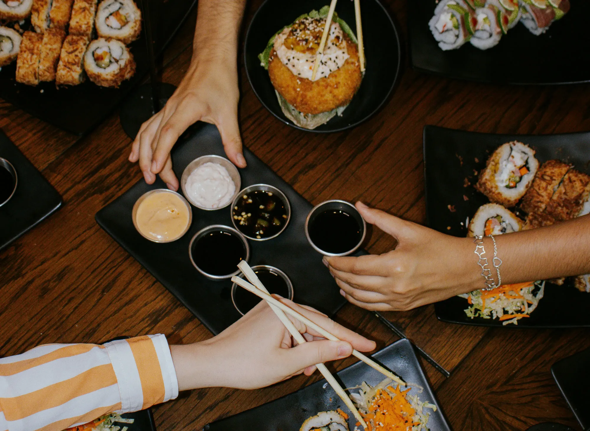 Hands reaching for sushi rolls and dipping sauces on a wooden table, with various sushi dishes and chopsticks visible.