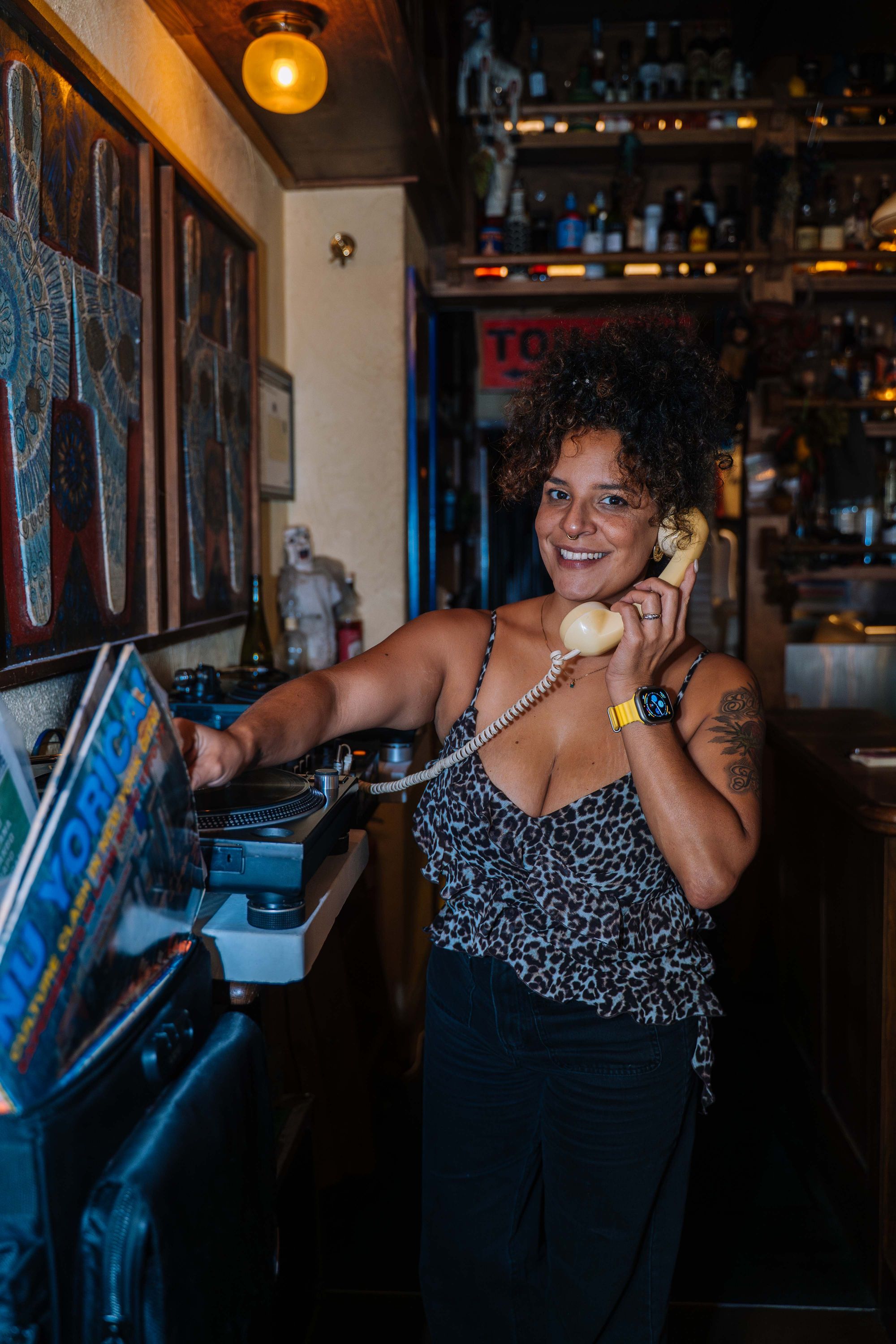 Person with curly hair in a leopard print top holds a vintage phone in a cozy bar, standing near a record player and smiling.
