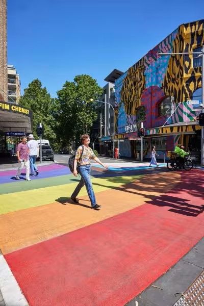 Person walking on a colourful rainbow crosswalk beneath a bright blue sky, with a vibrant mural on a nearby building in the background.