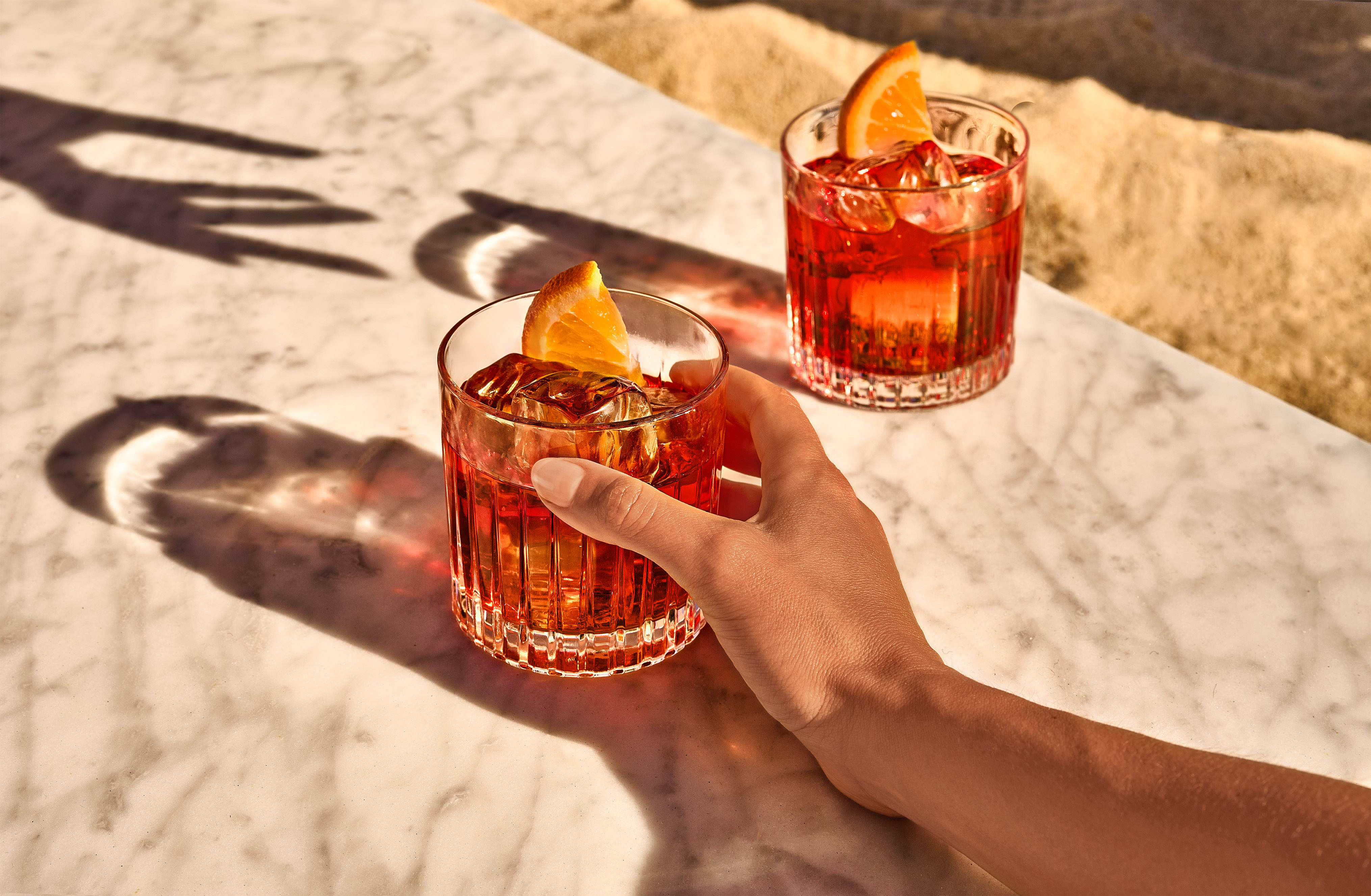 A hand holding a glass of red cocktail with an orange slice, next to another similar glass on a marble table in sunlight.