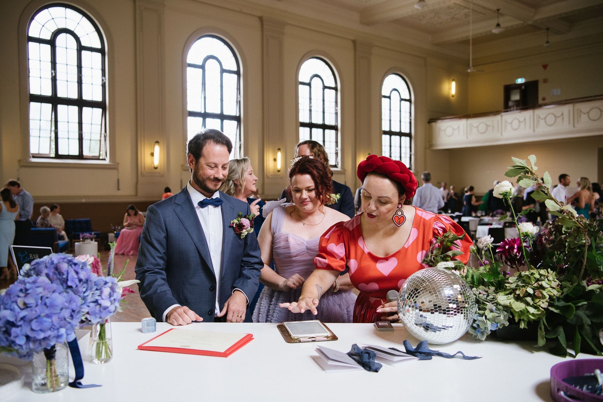 People signing documents at a formal event in a decorated hall, with flowers, a disco ball, and large arched windows in the background.