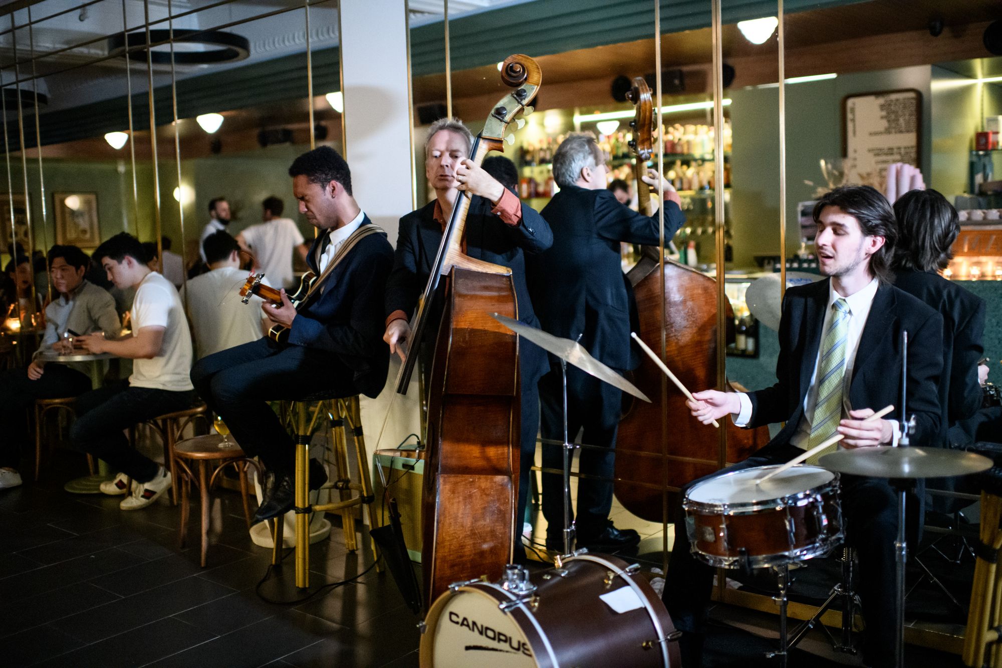 A jazz band performs in a lively bar, featuring a guitarist, double bass players, and a drummer, with patrons seated nearby.