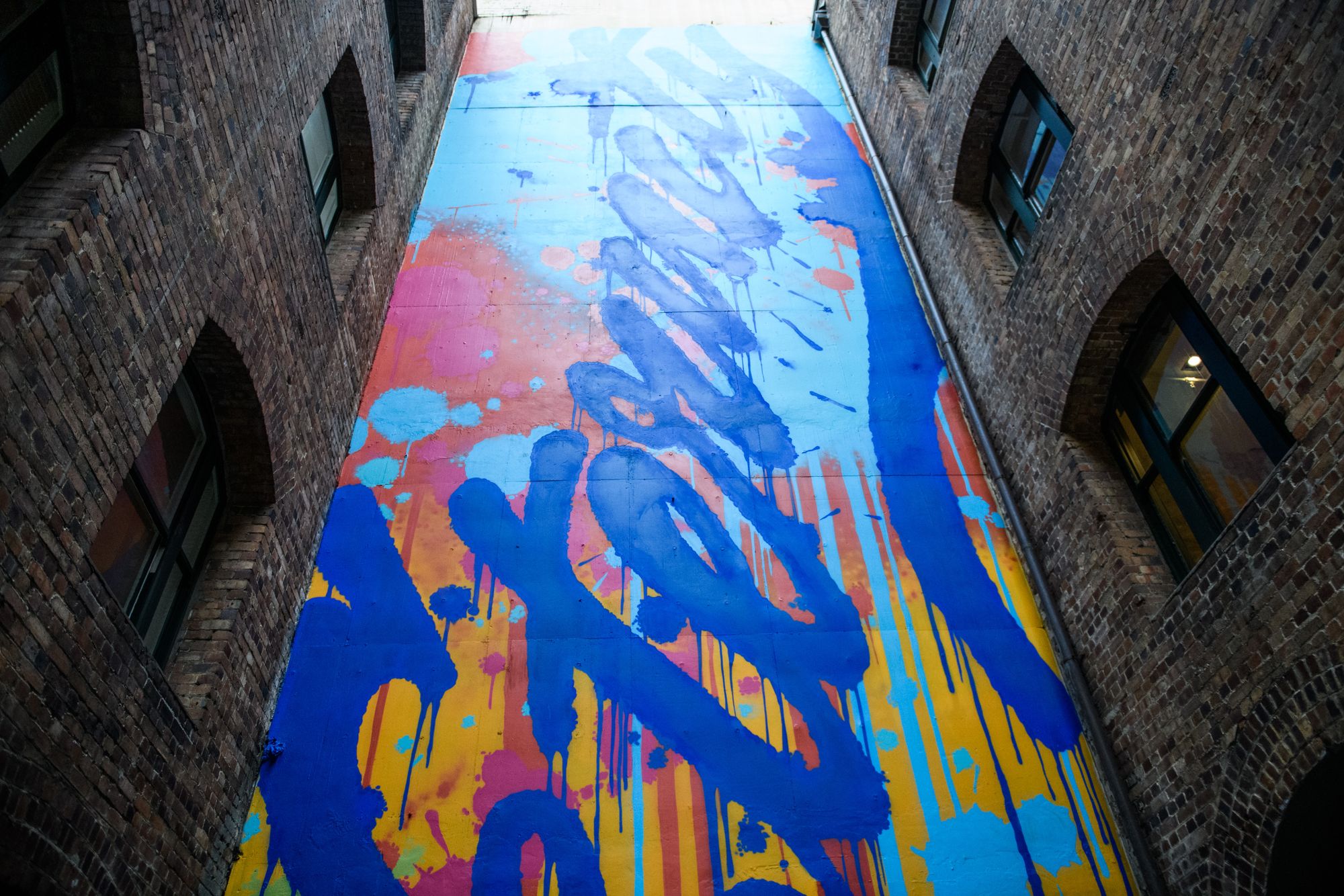Colorful graffiti with blue and orange splashes covers a tall wall between brick buildings, viewed from below.