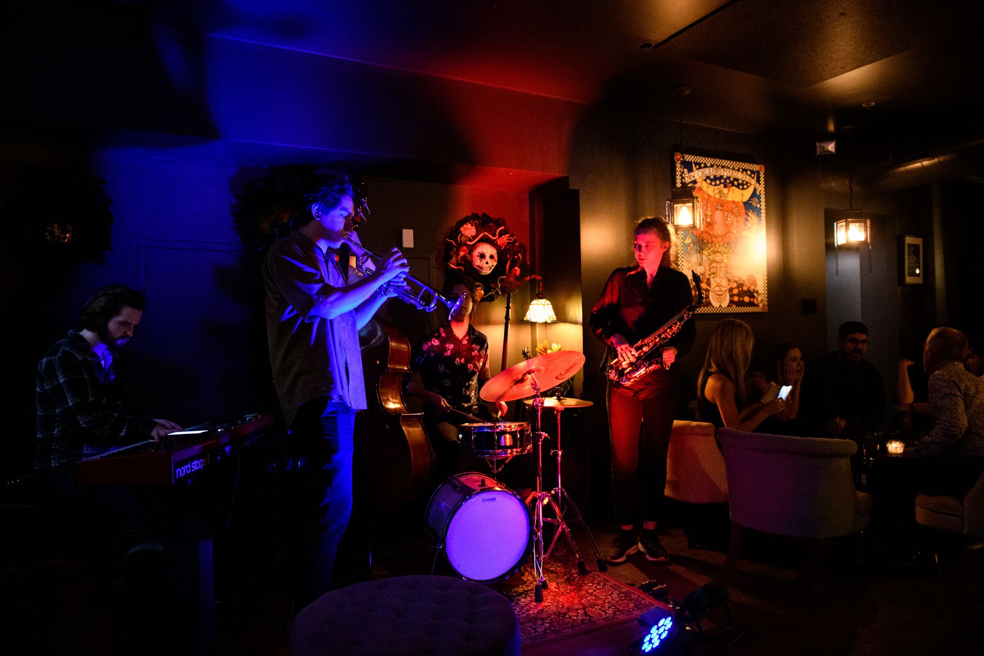 A jazz band performs on a dimly lit stage, featuring a keyboardist, trumpeter, bassist, drummer, and saxophonist, with an audience seated nearby.