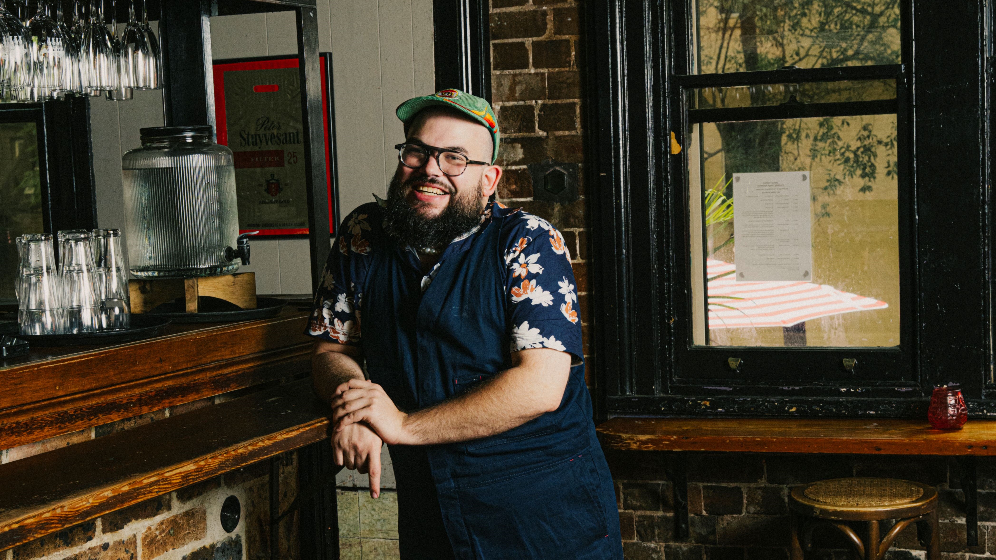 Smiling man in floral shirt and cap leans on a wooden bar in a cozy room with brick walls and a window.