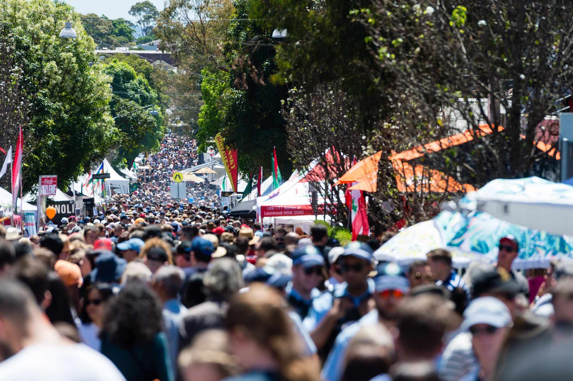 A bustling outdoor market with a large crowd, tents, and colorful flags lining a tree-lined street under a clear sky.