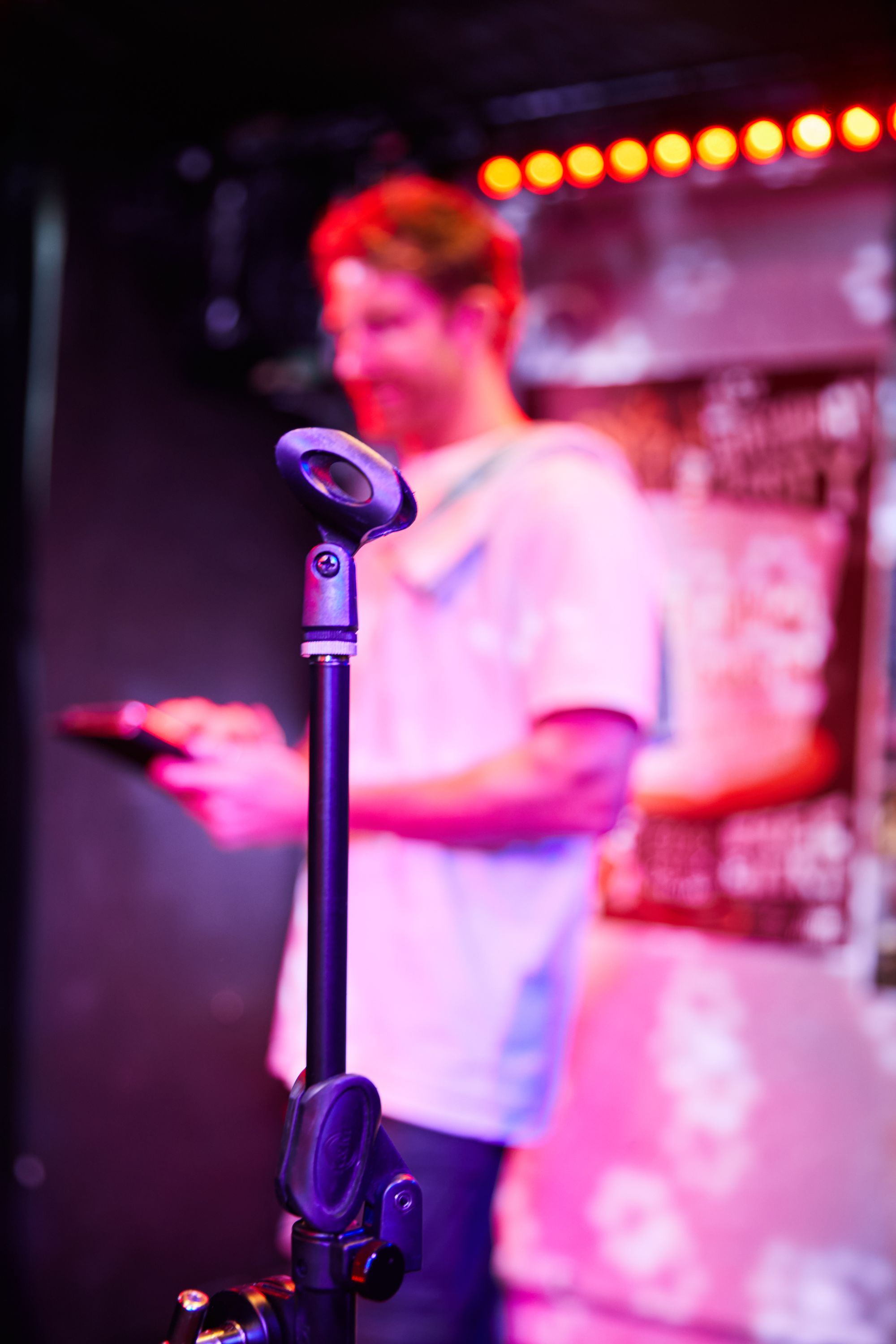 Blurred man in a white shirt holding a tablet, standing behind a focused microphone stand on a dimly lit stage.