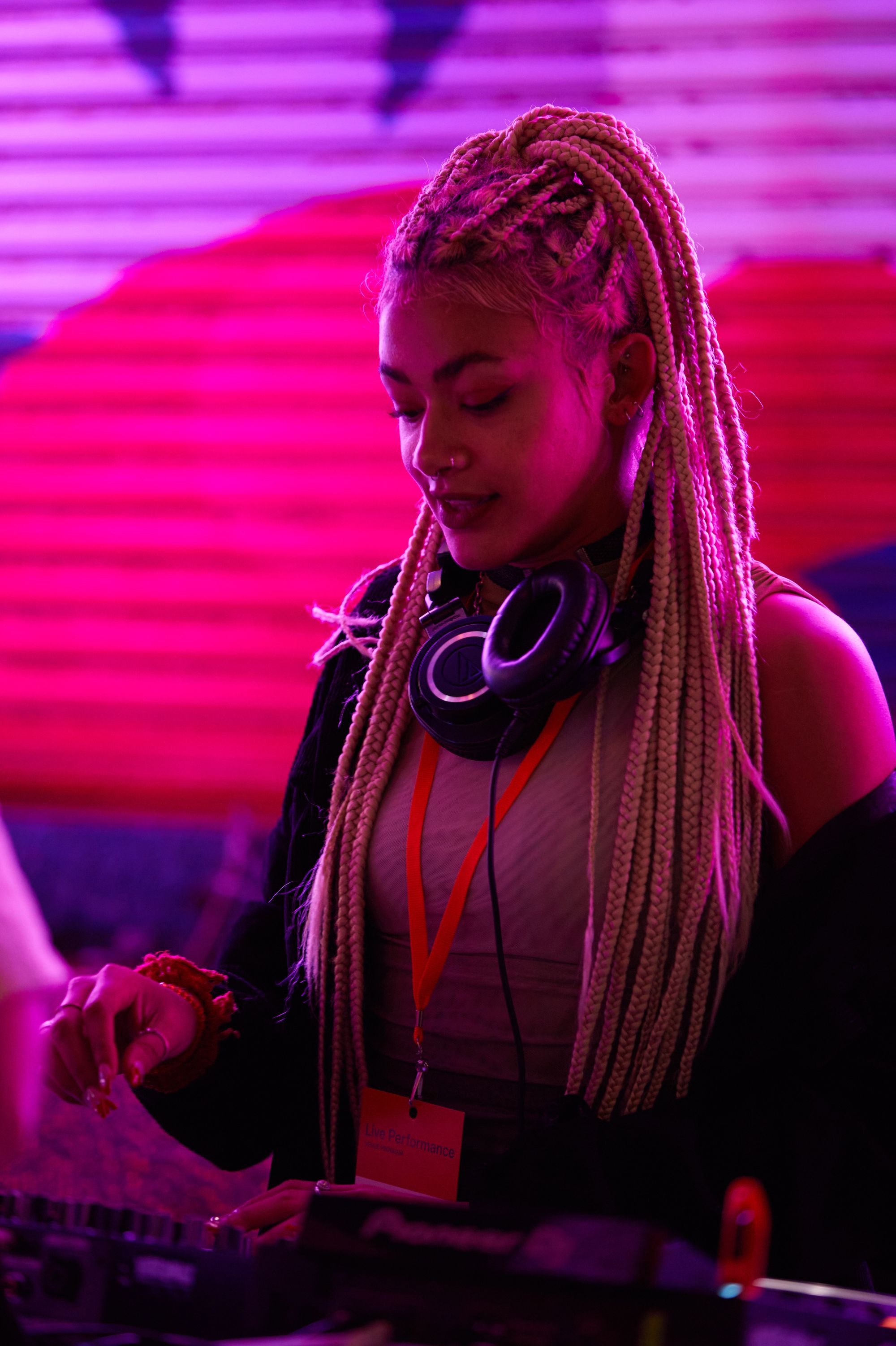 DJ with braided hair and headphones mixes music at a vibrant, pink-lit venue.