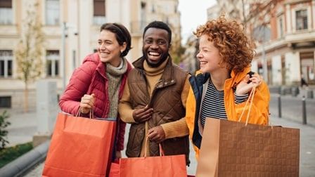 Three people happily carrying shopping bags on a city street, dressed warmly in jackets, with buildings in the background.