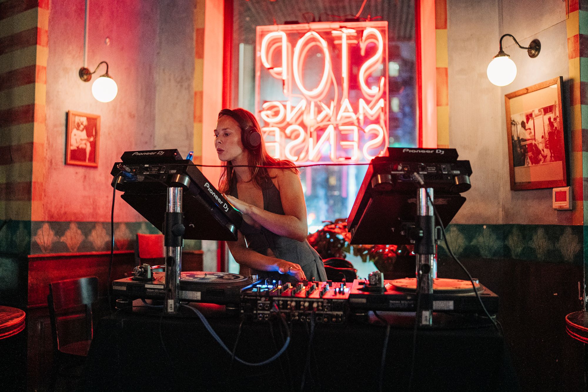DJ performing in a dimly lit room with a neon sign reading "STOP MAKING SENSE" in the background, surrounded by vintage decor.